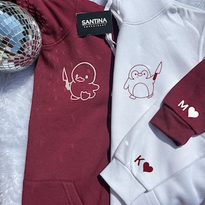 Puede incluir: Se muestran dos sudaderas con capucha, una burdeos y otra blanca. La sudadera burdeos presenta un pato de dibujos animados blanco con un cuchillo, mientras que la sudadera blanca tiene un pingüino rojo con un cuchillo. Las mangas muestran iniciales y corazones.