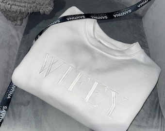 Puede incluir: Sudadera blanca con la palabra "WIFEY" bordada en blanco en la parte delantera. La sudadera está doblada y sobre una superficie gris.