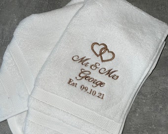 Könnte beinhalten: Zwei weiße Handtücher mit gestickten braunen Herzen und dem Text "Mr & Mrs George Est 09.10.21".