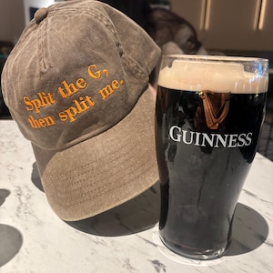 May include: A brown baseball cap with the text "Split the G, then split me" embroidered in orange. Beside the cap is a pint glass of Guinness stout with the Guinness logo.