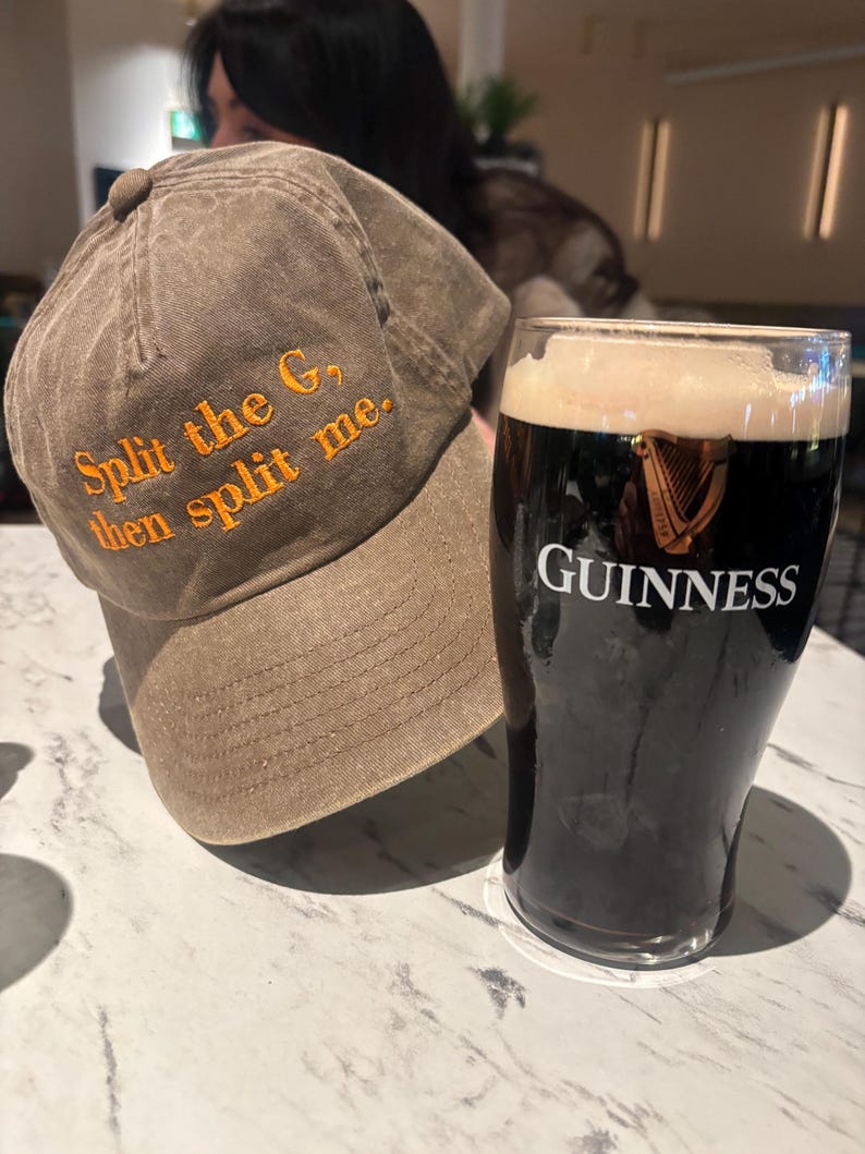May include: A brown baseball cap with the text "Split the G, then split me" embroidered in orange. Beside the cap is a pint glass of Guinness stout with the Guinness logo.