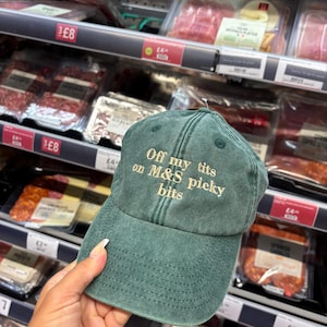 Puede incluir: Gorra de béisbol verde con el texto bordado "Off my tits on M&S picky bits". La gorra se muestra frente a un estante de supermercado con alimentos envasados.