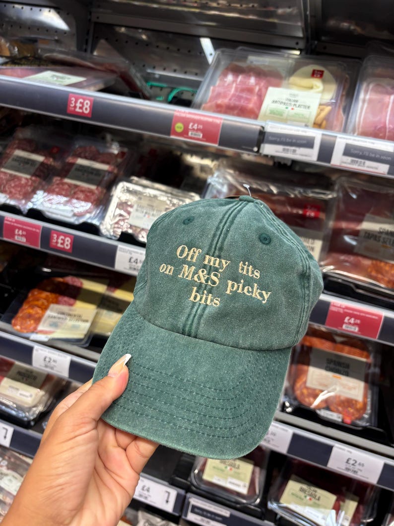 May include: A green baseball cap with the text "Off my tits on M&S picky bits" embroidered in a lighter color. The cap is held in front of a grocery store shelf with various packaged food items.