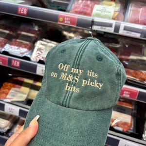 May include: A green baseball cap with the text "Off my tits on M&S picky bits" embroidered in a light tan color. The cap is made of a soft, worn material and has a curved brim.