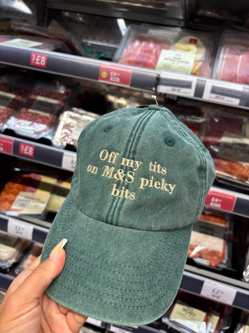 May include: A green baseball cap with the text "Off my tits on M&S picky bits" embroidered in a light tan color. The cap is made of a soft, worn material and has a curved brim.