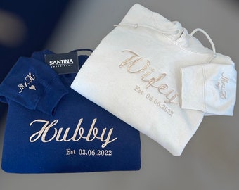 May include: Two hooded sweatshirts, one navy blue and one white, with embroidered text. The navy blue sweatshirt has "Hubby Est 03.06.2022" on the front and "Mrs &" on the sleeve. The white sweatshirt has "Wifey Est 03.06.2022" on the front and "Mrs &" on the sleeve.