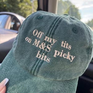 May include: A dark green baseball cap with the embroidered text "Off my tits on M&S picky bits" in a light beige color. The cap is made of a textured fabric and has a curved brim.