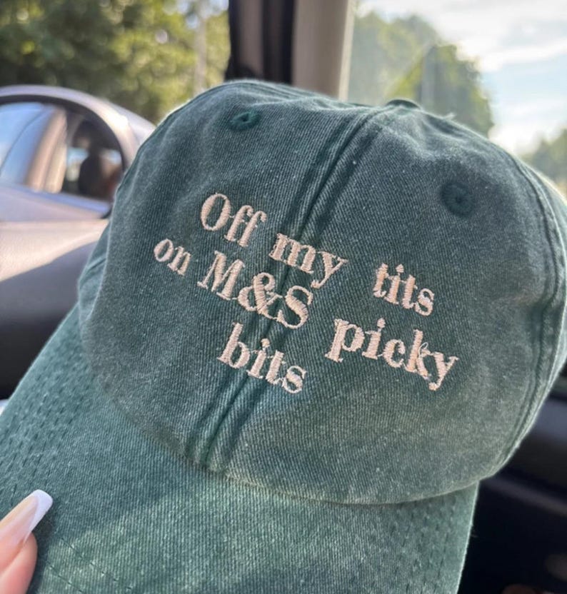 May include: A dark green baseball cap with the embroidered text "Off my tits on M&S picky bits" in a light beige color. The cap is made of a textured fabric and has a curved brim.