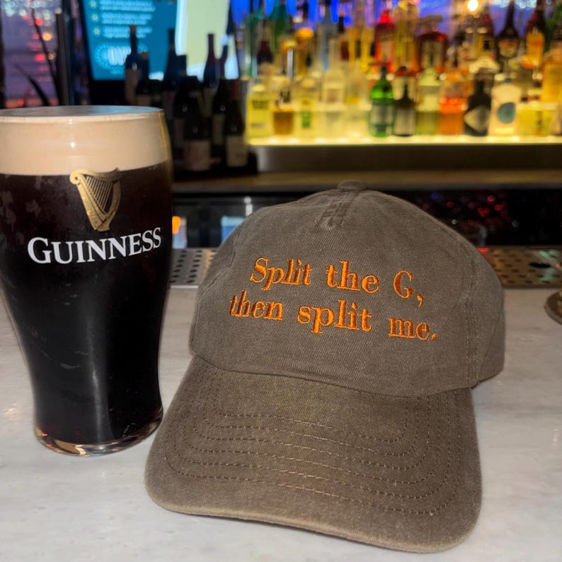 May include: A pint of Guinness stout next to a brown baseball cap. The cap has orange embroidered text that reads "Split the G, then split me."