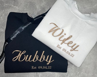 May include: Two black and white sweatshirts with gold embroidery. The black sweatshirt says "Hubby Est 09.04.22" and the white sweatshirt says "Wifey Est 09.04.22".