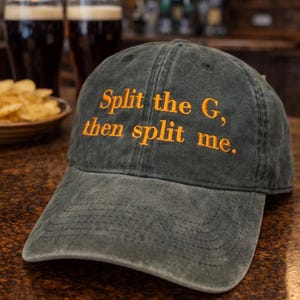 Op de afbeelding: Een donkergroene baseball cap met de tekst "Split the G, then split me" geborduurd in oranje. De cap ligt op een bruin oppervlak, met twee glazen donker bier en een schaal chips op de achtergrond.