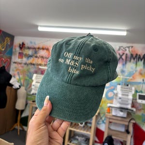 May include: A dark green baseball cap with the embroidered text "Off my tits on M&S picky bits" in white. The cap is held up by a hand. The background is a colorful wall with graffiti and sewing machines.