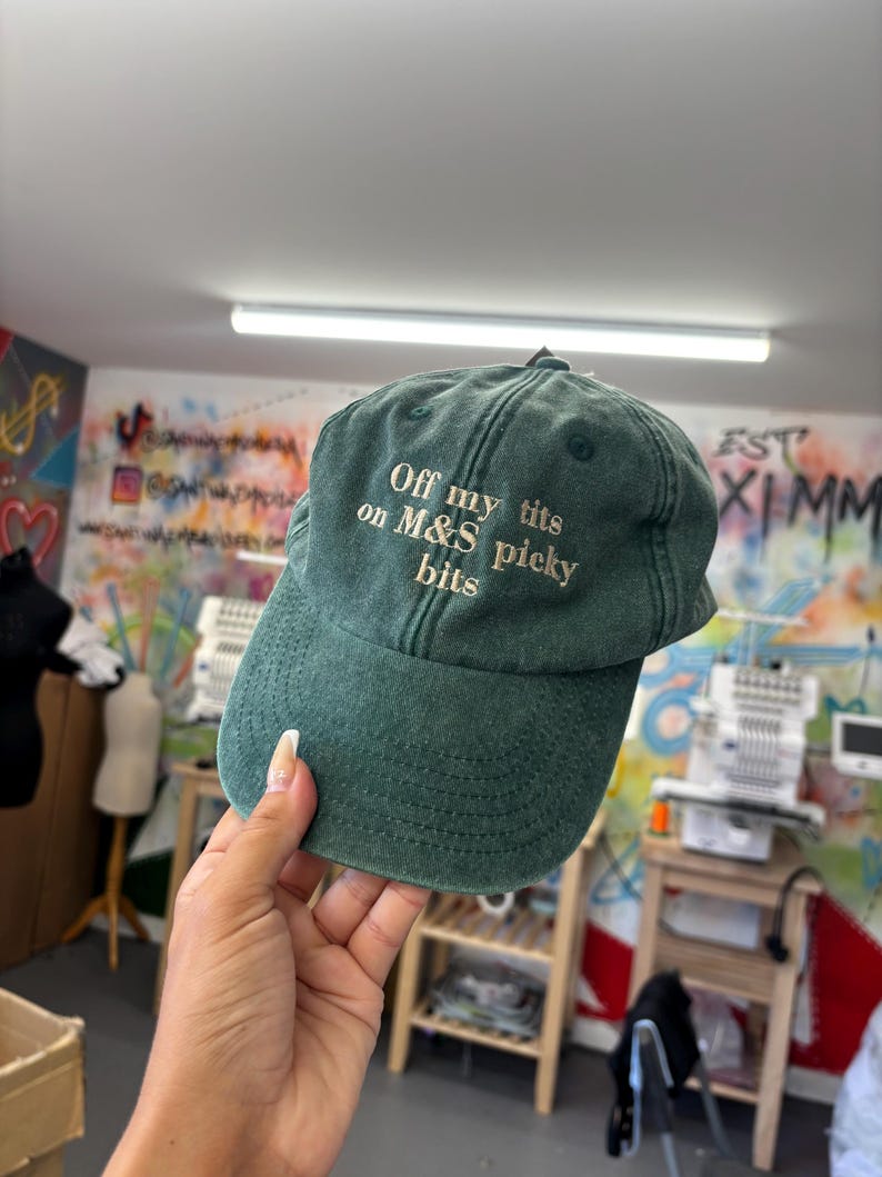 May include: A dark green baseball cap with the embroidered text "Off my tits on M&S picky bits" in white. The cap is held up by a hand. The background is a colorful wall with graffiti and sewing machines.