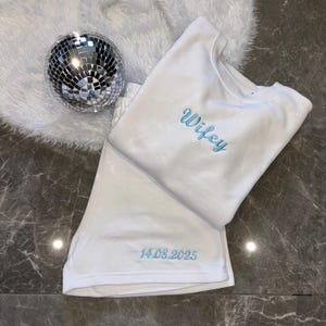 Puede incluir: Pijama de algod&oacute;n blanco con la palabra "wifey" bordada en azul en la parte superior y la fecha "14.08.2025" bordada en azul en la parte inferior.