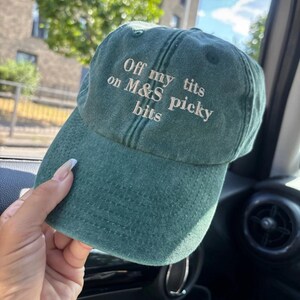 May include: A green baseball cap with the text "Off my tits on M&S picky bits" embroidered in white. The cap has a curved brim and a slightly distressed appearance, suggesting a vintage or worn style.