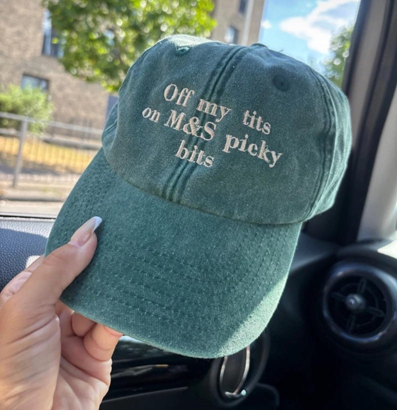 May include: A green baseball cap with the text "Off my tits on M&S picky bits" embroidered in white. The cap has a curved brim and a slightly distressed appearance, suggesting a vintage or worn style.