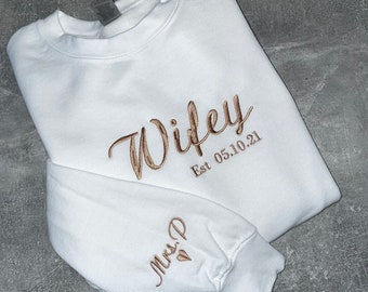 Puede incluir: Sudadera blanca con el texto "Wife" en cursiva con "Est 05.10.21" debajo. La manga izquierda tiene el texto "Mrs. D" con un corazón.