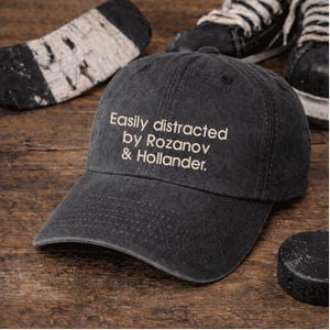 Peut inclure: Casquette de baseball gris foncé avec le texte brodé "Easily distracted by Rozanov & Hollander." La casquette est posée sur une surface en bois, avec une rondelle de hockey, une crosse de hockey et une paire de patins de hockey en arrière-plan.