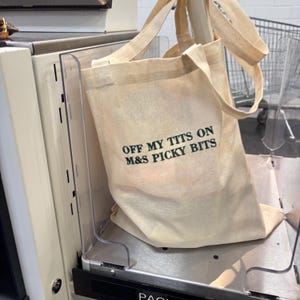 Könnte beinhalten: Beige Canvas-Tragetasche mit dem Text "OFF MY TITS ON M&S PICKY BITS" in grünen Buchstaben. Die Tasche hat zwei Griffe und steht auf einer Metalloberfläche.