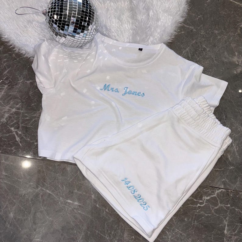 Puede incluir: Conjunto de pijama de algod&oacute;n blanco con bordado azul. La parte superior dice "Mrs. Jones" y los pantalones cortos dicen "14.08.2025".