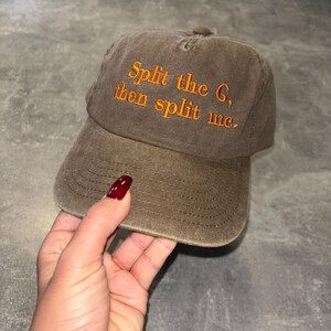 May include: A brown baseball cap with the text "Split the G, then split me" embroidered in orange. The cap has a curved brim and a fabric strap closure. The hat is made of a soft, worn material.
