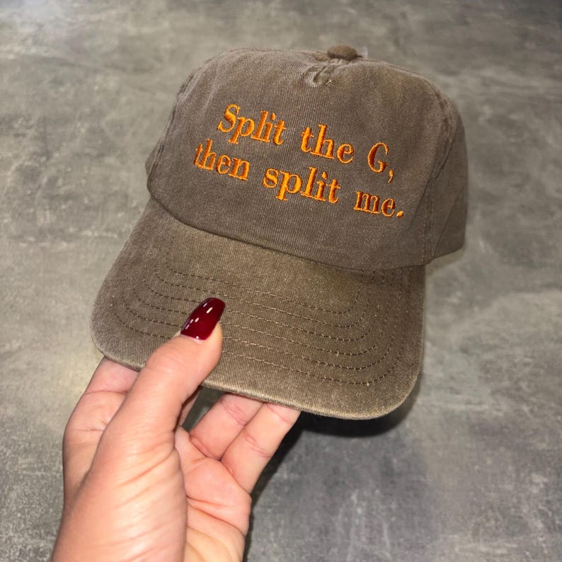 May include: A brown baseball cap with the text "Split the G, then split me" embroidered in orange. The cap has a curved brim and a fabric strap closure. The hat is made of a soft, worn material.