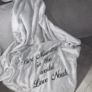 Puede incluir: Una manta de forro polar gris con el texto "Best Mummy in the world Love Noah" bordado en negro.