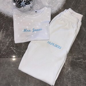 Puede incluir: Camiseta blanca con un patr&oacute;n de diamantes y el texto "Mrs. Jones" bordado en azul. Pantal&oacute;n de ch&aacute;ndal blanco con la fecha "14.08.2025" bordada en azul.