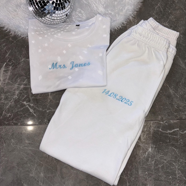 Puede incluir: Camiseta blanca con un patr&oacute;n de diamantes y el texto "Mrs. Jones" bordado en azul. Pantal&oacute;n de ch&aacute;ndal blanco con la fecha "14.08.2025" bordada en azul.