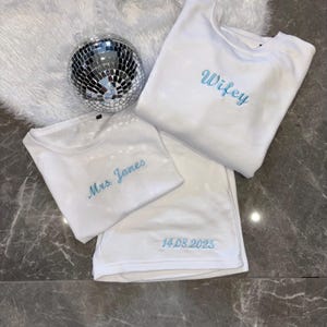 Puede incluir: Tres camisetas blancas con bordados azules. La camiseta de arriba dice "Wifey". La camiseta del medio dice "Mrs. James". La camiseta de abajo dice "14.08.2025".