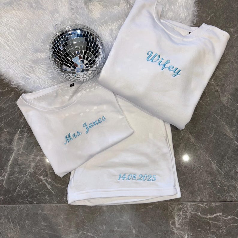 Puede incluir: Tres camisetas blancas con bordados azules. La camiseta de arriba dice "Wifey". La camiseta del medio dice "Mrs. James". La camiseta de abajo dice "14.08.2025".
