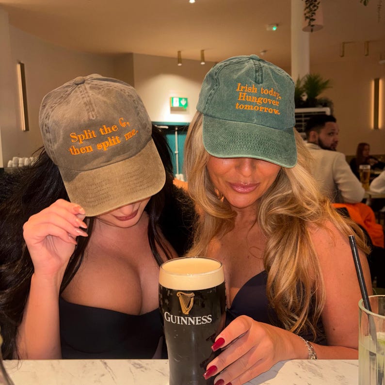 May include: Two baseball caps with text, one brown and one green, are worn by two people. The brown cap says "Split the G, then split me" in orange. The green cap says "Irish today, Hungover tomorrow" in orange. A pint of Guinness is in the foreground.
