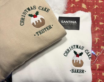 May include: Two Christmas-themed sweatshirts, one beige and one white, with embroidered Christmas cake designs. The beige sweatshirt has the text "CHRISTMAS CAKE. TESTER." The white sweatshirt has the text "CHRISTMAS CAKE. BAKER."