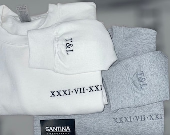 May include: Two sweatshirts, one white and one gray, with the same embroidered design. The design includes the initials "T&L" and the Roman numeral date "XXXI VII-XXI". The sweatshirts are stacked on top of a black tag that reads "SANTINA EMBROIDERY".