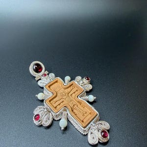 Puede incluir: Colgante de plata con forma de cruz, con un elemento central de madera con una figura religiosa grabada. La cruz est&aacute; adornada con piedras preciosas rojas y perlas blancas, con intrincados detalles de filigrana de plata.