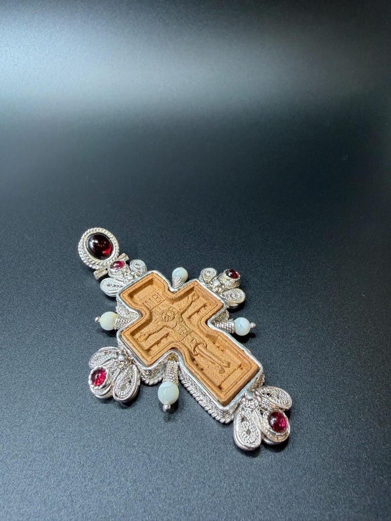 Puede incluir: Colgante de plata con forma de cruz, con un elemento central de madera con una figura religiosa grabada. La cruz est&aacute; adornada con piedras preciosas rojas y perlas blancas, con intrincados detalles de filigrana de plata.