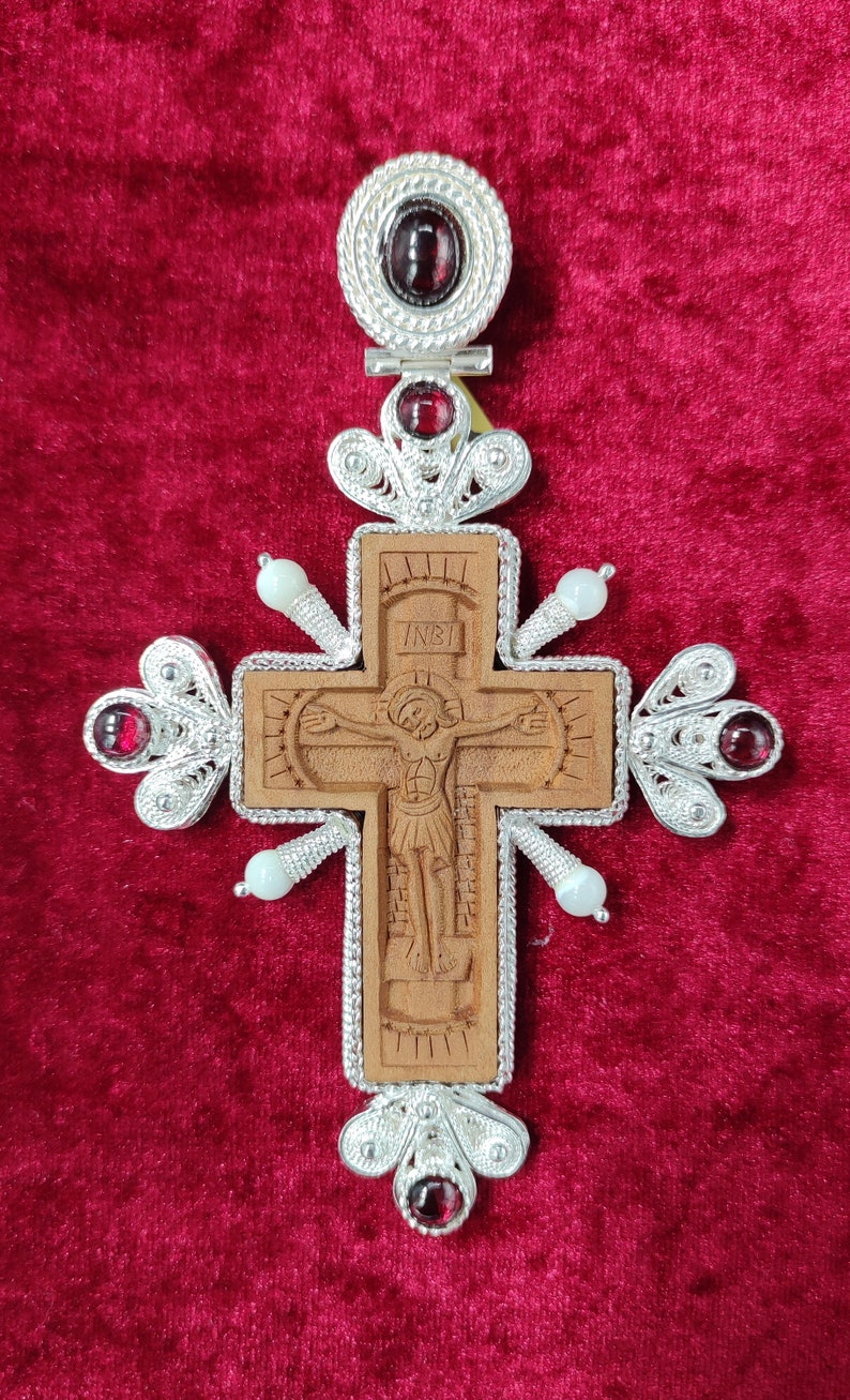 Puede incluir: Un colgante de cruz de madera detallado con una imagen tallada de Jesucristo. La cruz est&aacute; enmarcada con filigrana de plata ornamentada y adornada con piedras preciosas rojas y cuentas blancas. El fondo es un terciopelo rojo oscuro.