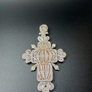 Puede incluir: Colgante de cruz de filigrana plateada con un intrincado dise&ntilde;o calado. La cruz presenta una secci&oacute;n central con un fondo texturizado marr&oacute;n claro y peque&ntilde;as cuentas verde p&aacute;lido. Una joya religiosa detallada.