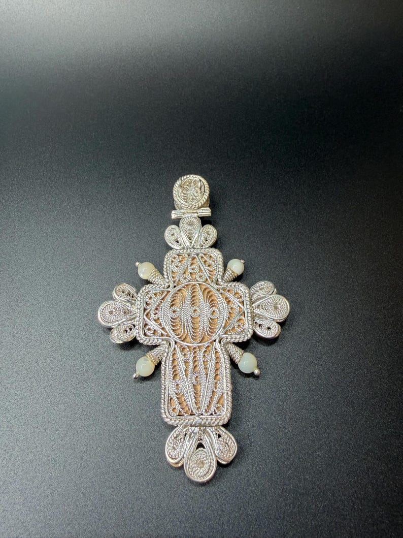 Puede incluir: Colgante de cruz de filigrana plateada con un intrincado dise&ntilde;o calado. La cruz presenta una secci&oacute;n central con un fondo texturizado marr&oacute;n claro y peque&ntilde;as cuentas verde p&aacute;lido. Una joya religiosa detallada.