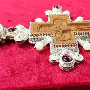 Puede incluir: Un colgante de cruz de plata detallado con un centro de madera tallada. La cruz presenta intrincados trabajos de filigrana, acentos de piedras preciosas rojas y peque&ntilde;as perlas blancas. El colgante se muestra sobre una superficie de terciopelo rojo.