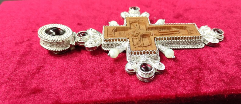 Puede incluir: Un colgante de cruz de plata detallado con un centro de madera tallada. La cruz presenta intrincados trabajos de filigrana, acentos de piedras preciosas rojas y peque&ntilde;as perlas blancas. El colgante se muestra sobre una superficie de terciopelo rojo.