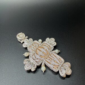 Puede incluir: Colgante de cruz de filigrana plateada con un intrincado dise&ntilde;o calado. La cruz presenta un elemento central texturizado y est&aacute; adornada con peque&ntilde;as cuentas redondas de color blanquecino. El colgante descansa sobre una superficie oscura, mostrando su detallada artesan&iacute;a.