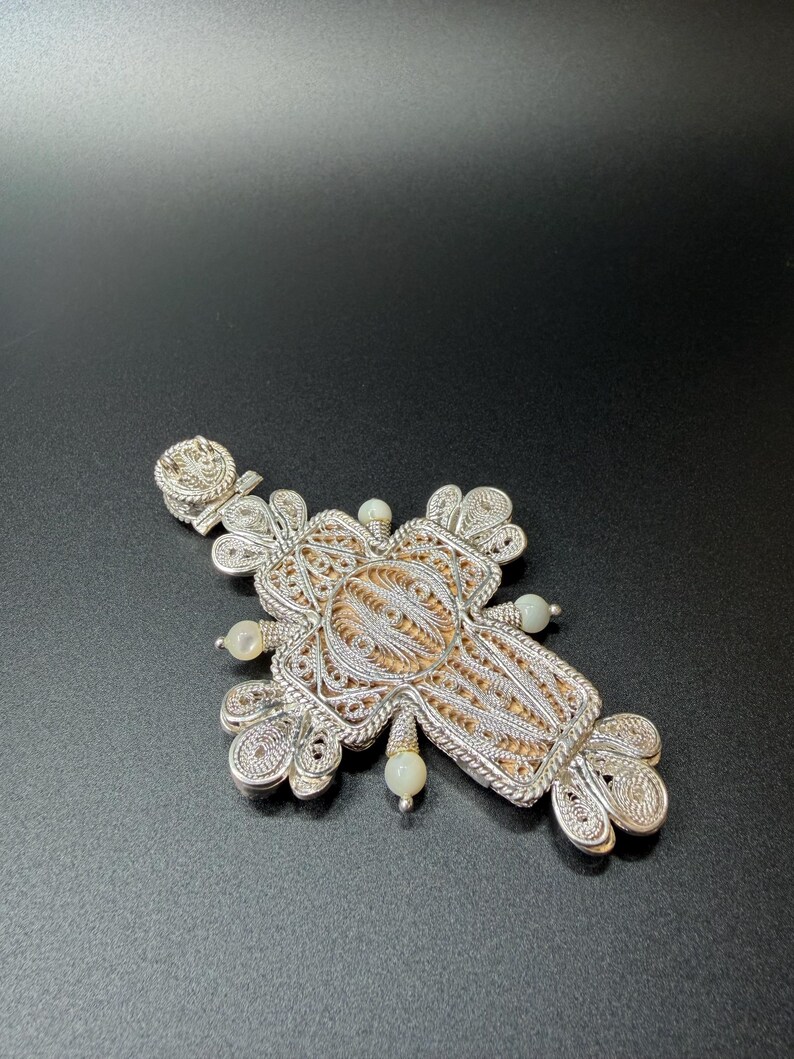 Puede incluir: Colgante de cruz de filigrana plateada con un intrincado dise&ntilde;o calado. La cruz presenta un elemento central texturizado y est&aacute; adornada con peque&ntilde;as cuentas redondas de color blanquecino. El colgante descansa sobre una superficie oscura, mostrando su detallada artesan&iacute;a.