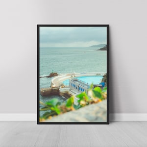 Puede incluir: Una impresión enmarcada que muestra una piscina costera con agua turquesa, con el mar de fondo y un cielo nublado. La piscina tiene un borde blanco y detalles azules. La impresión está enmarcada en negro.