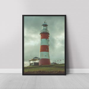 Puede incluir: Impresión enmarcada de un faro con rayas rojas y blancas. El faro se alza contra un cielo nublado, con un pequeño edificio en su base y banderas a lo lejos. La imagen está enmarcada en un marco negro.