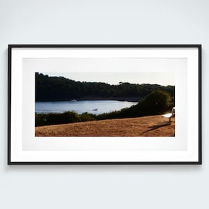 May include: A framed landscape photograph featuring a golden-brown grassy hill in the foreground, a calm blue bay with a small boat in the middle ground, and a dark green forested coastline under a bright sky, finished with a black frame.