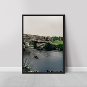 Puede incluir: Impresión enmarcada de un puente de piedra sobre un río. El puente, de piedra gris desgastada, tiene una abertura arqueada. El agua oscura refleja el cielo nublado. El follaje verde bordea las orillas. Un paisaje tranquilo.