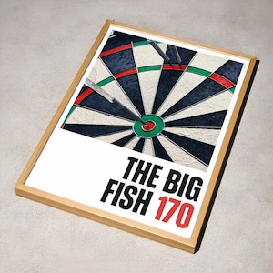 Peut inclure: Impression encadrée d'une cible de fléchettes avec trois fléchettes plantées. La cible présente des sections noires, blanches, rouges et vertes. Le texte "THE BIG FISH 170" est imprimé en noir et rouge en dessous.