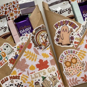 May include: Two brown paper gift cones filled with Cadbury Dairy Milk and Galaxy chocolate bars, decorated with autumn leaf patterned paper and various ghost-themed stickers featuring text like "SPOOKY VIBE," "LET'S GO GHOULS," and "READ MORE BOOKS."