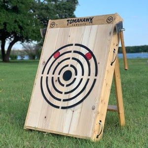 May include: Wooden axe throwing target with a black and white bullseye and red scoring rings. The target is mounted on a wooden A-frame stand. The text "TOMAHAWK TARGETS" is printed on the target.